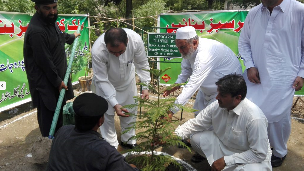 4 men in white and two in black plant a tree with shovel, green signs in back