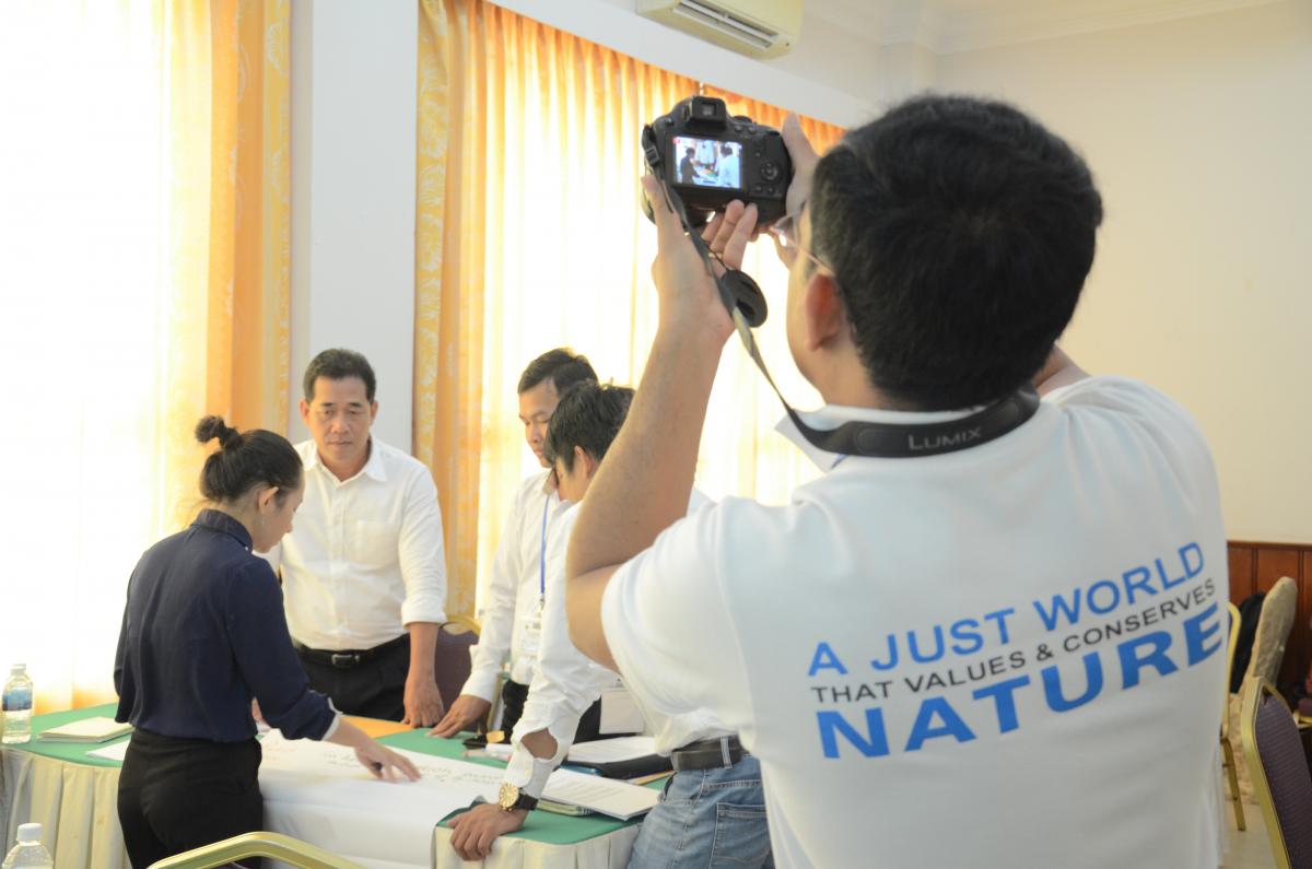 A man in a t-shirt that reads 'A just world that values and conserves nature' takes a photo of workshop participants