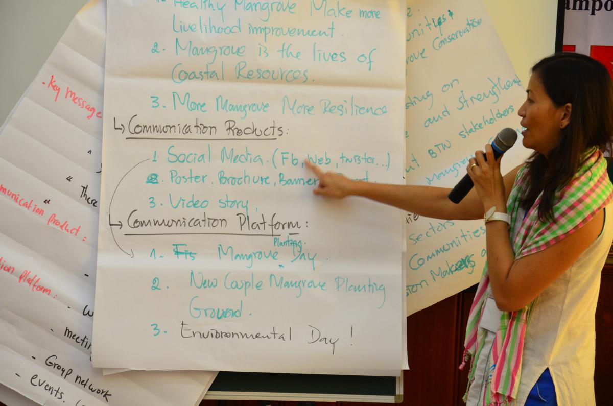 A woman holding a microphone points at a large pad of presentation paper with green writing