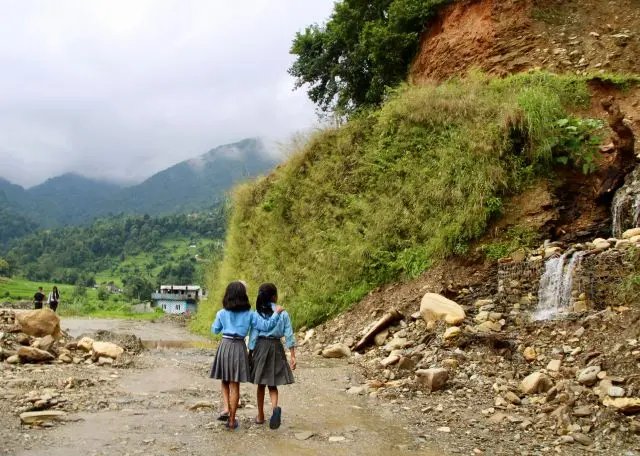 Two young children in blue and grey clothing walk along a road, with greenery to their right.