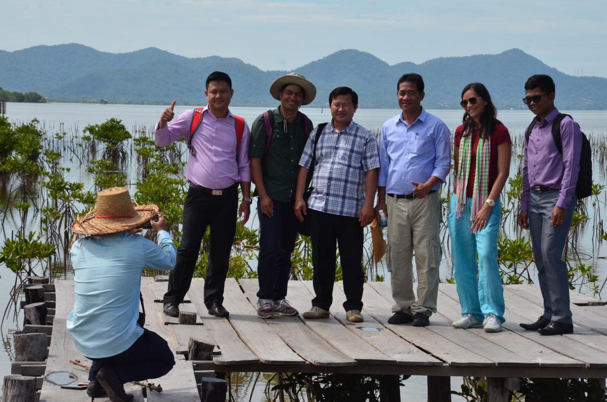 Six people stand in front of mangroves and mountains in the distance while another takes their photo