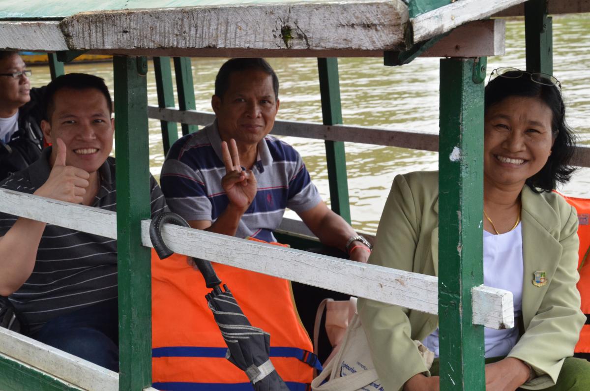 Two men and a woman in a boat smile at the camera and give thumbs up and peace signs