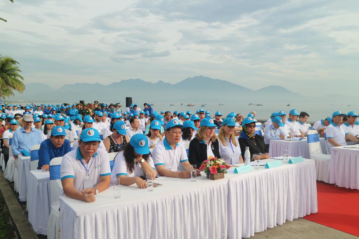 Hundreds of people in blue hats sit at rows of tables, with the sea and distant mountains in the background