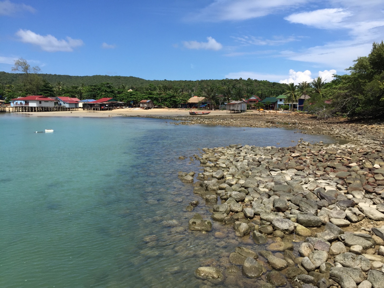 A rocky beach, clear blue water, several small houses on stilts in the background, and short island forest beyond